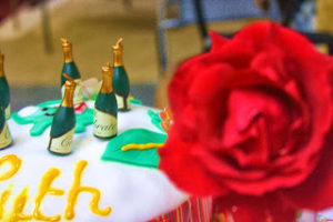 A red rose fills the right hand side of the frame and on the left, behind the rose, can be seen the top of a cake with small champagne bottle candles on white and green frosting.