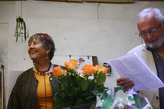 Snapshot of Ruth Hartley in a yellow dress, laughing as she looks  to her right, while her partner John Corley reads or sings from a sheet he holds in front of him. They stand behind a vase of yellow-orange roses.  