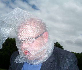 Head and shoulders shot of a bespectacled man in a dark jacket and a blue shirt with his head entirely covered in bubble wrap, except for his lips, which are visible through a hole in the wrap. Against a cloudy sky, he bends his head slightly forward.
