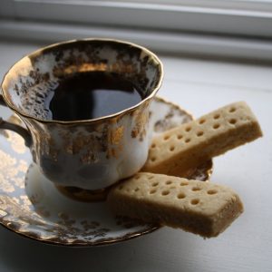 A beautiful and delicate cup and saucer are in the centre of the frame. the cup is full of dark coffee and there are two fingers of shortbread on the saucer. 